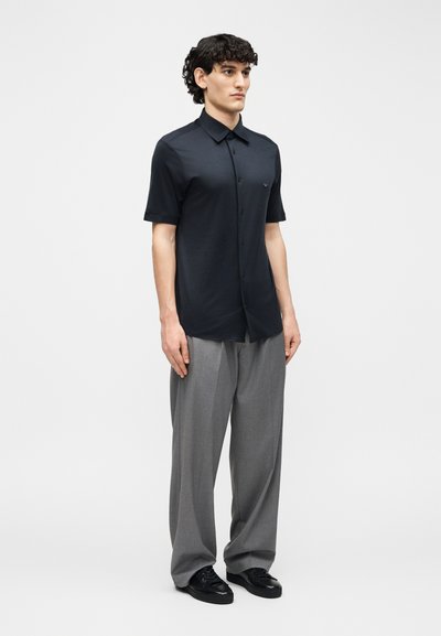 Άνδρας που στέκεται φορώντας μαύρο πουκάμισο με κοντά μανίκια και κουμπιά, γκρι παντελόνι φαρδιάς γραμμής και μαύρα παπούτσια, μπροστά από απλό φόντο.