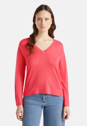 Korallfarbener V-Ausschnitt-Pullover aus weichem Strickmaterial. Lange Ärmel, entspannter Schnitt, dezenter gerippter Saum. Kombiniert mit hellblauen Jeans.