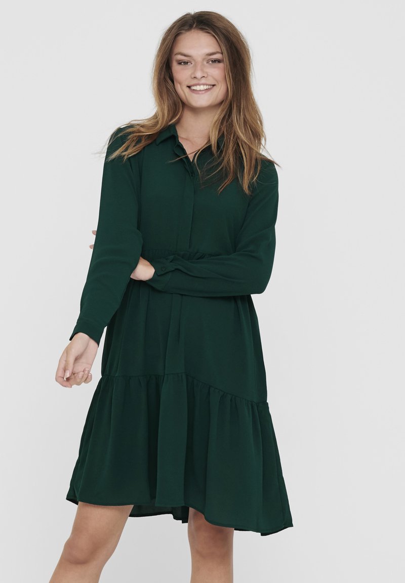 Robe verte à manches longues avec col, taille cintrée et ourlet à volants superposés, fabriquée en tissu doux. Texture lisse et design décontracté.