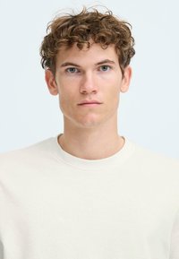 Light beige crew-neck t-shirt made of cotton, featuring a smooth texture and short sleeves, worn by a person with curly hair and blue eyes.
