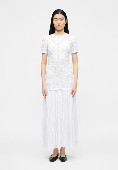 Žena s dlouhými černými vlasy stojí a má na sobě bílý texturovaný krátký rukáv a plisovanou dlouhou sukni, doplněné černými balerínami na bílém pozadí.