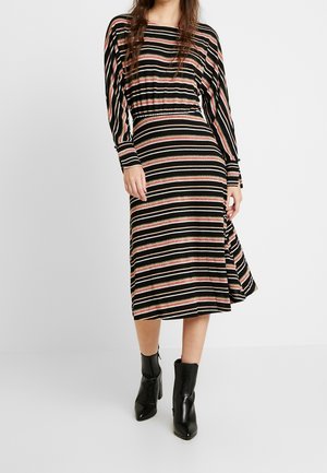 Kvinde iført en midi-kjole med sorte, beige og pink horisontale striber med lange ærmer og sorte ankelstøvler, stående mod en almindelig baggrund.