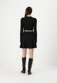 Black long-sleeve dress with a fitted bodice and flared skirt, featuring a branded waistband. Black knee-high boots with a slight heel.