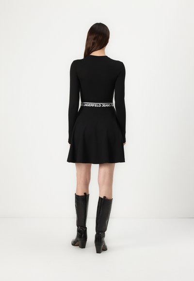 Robe noire à manches longues avec un corsage ajusté et une jupe évasée, dotée d'une ceinture de marque. Bottes noires montantes avec un léger talon.