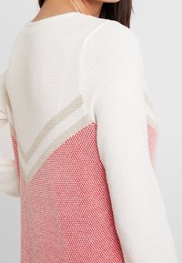 Pull blanc en tricot avec un motif en forme de V beige et rouge. Tissu texturé avec un motif contrastant rouge et crème.