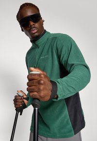 Green fleece pullover with a half-zip collar, black accents, and a soft texture. Model holds trekking poles, wearing large dark sunglasses.