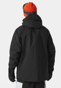 Chaqueta negra impermeable con capucha, que presenta una textura suave, puños elásticos y una insignia en el hombro izquierdo. Llevada con un gorro naranja.