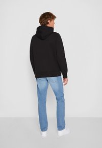 Felpa nera con orlo e polsini a coste, indossata con jeans azzurri chiari e sneakers bianche, vista da dietro su uno sfondo neutro.