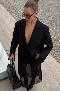 Moteris su sukulniu ir dideliais akiniais nuo saulės, vilkinti juodą švarką ir permatomą sijoną, laiko juodą rankinę ir stovi ant akmeninio grindinio.