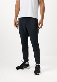 Schwarze Sporthose mit Reißverschlusstaschen, strukturierten unteren Paneelen und eng anliegenden Knöchelabschlüssen. Getragen mit schwarzen Turnschuhen mit einem weißen Logo.
