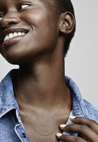 Zlati verižni nakit, nosen čez denim srajco, ki poudarja vratni izrez in ovratnik. Gladka tekstura kože in nevtralni lak za nohte sta vidna.