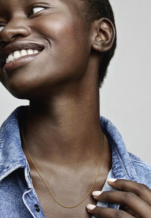 Zlatý retiazkový náhrdelník nosený cez džínsovú košeľu, zdôrazňujúci výstrih a golier. Hladká textúra pleti a neviditeľný lak na nechtoch.