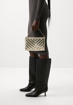 Złota pikowana torebka z wzorem w jodełkę i łańcuszkowym paskiem, trzymana w jednej ręce. Modelka ubrana w prążkowaną, dopasowaną szarą sukienkę i czarne kozaki sięgające do kolan.