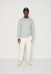 Lys grå sweatshirt, cremefarvede joggers med et grønt logo og hvide sneakers. Outfittet har en afslappet pasform og en subtil tekstur.