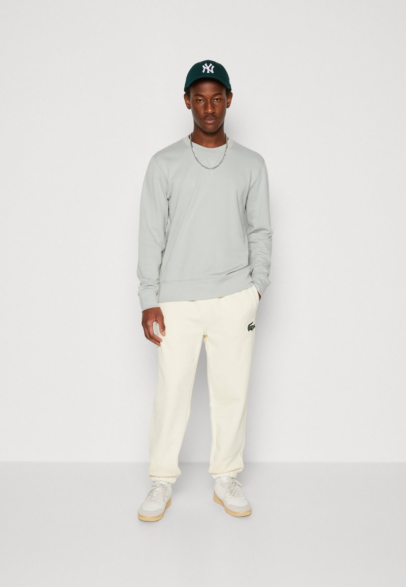 Lys grå sweatshirt, cremefarvede joggers med et grønt logo og hvide sneakers. Outfittet har en afslappet pasform og en subtil tekstur.