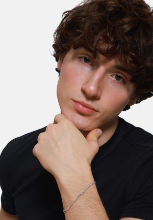 Braccialetto d'argento con design intrecciato indossato al polso. Il modello ha i capelli ricci e indossa una camicia nera, con la mano poggiata sul mento.