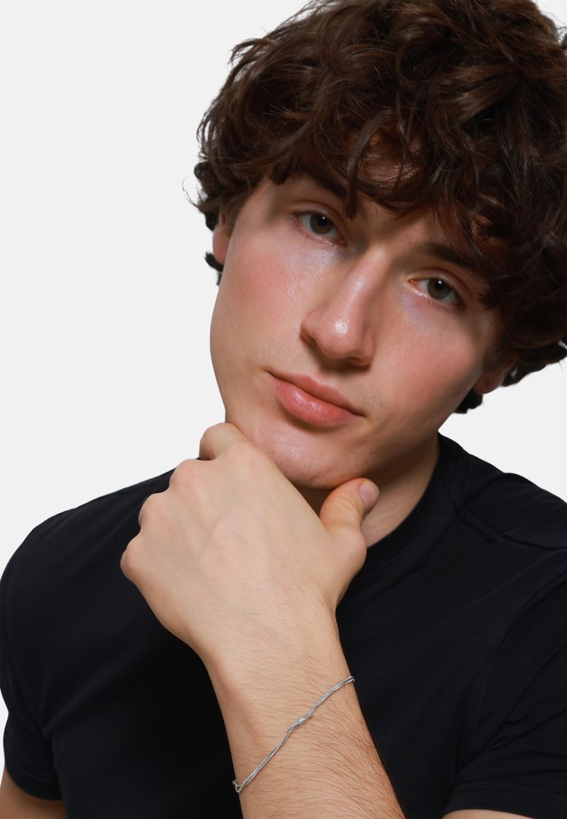 Silver bracelet with twisted design worn on the wrist. The model has curly hair and wears a black shirt, with their hand resting on their chin.