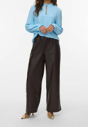 Vrouw draagt een lichtblauwe blouse met lange mouwen en een keyhole-halslijn, wijde donkerbruine leren broek en hakken met slangenprint.