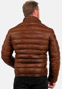 Man in een bruin gewatteerd leren jack, staande met zijn rug naar de camera tegen een eenvoudige witte achtergrond.