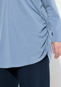 Maglia a maniche lunghe di un blu chiaro con dettagli arricciati ai lati, texture morbida e orlo curvo, abbinata a pantaloni blu navy scuri.