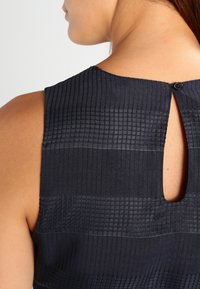 Ärm-lös marinblå topp med texturerad randig mönster, med en liten knappstängning på baksidan och ett nyckelhål detaljer.