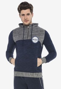 Navy- und graue Zip-Hoodie mit strukturiertem Stoff, Kängurutasche und Logo-Patch auf der Brust. Verstellbare Kordelzugkapuze.
