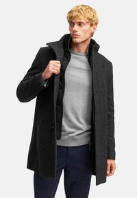 Dunkelgrauer Fischgrätenmantel mit hohem Kragen, Knopfverschluss und einem Reißverschluss-Innenfutter, getragen über einem grauen Pullover und marineblauen Hosen.