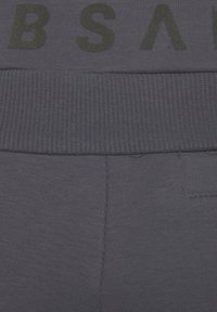 Dunkelgrauer Stoff mit einem gerippten Taillenbund zeigt große, schwarze Schriftzüge. Die Textur ist weich mit sichtbaren Nahtdetails.