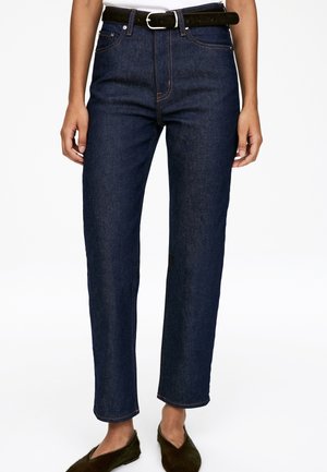 Dunkelblaue Jeans mit geradem Bein, ausgestattet mit orangefarbenen Nähten und fünf Taschen. Kombiniert mit einem schwarzen Gürtel und strukturiertem grünen Schuhwerk.