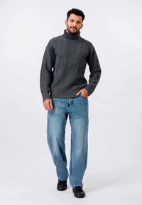 Pull en maille gris à col roulé avec des motifs en losange texturés, associé à un jean bleu clair et des chaussures noires. Se tenant devant un fond blanc.