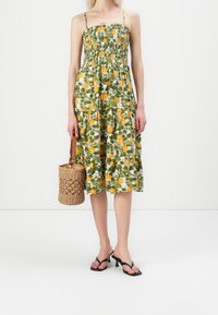 Robe midi florale avec un fond vert et des motifs de fruits jaunes et oranges, corsage froncé et fines bretelles. Accessoire : sac en osier.