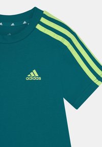 Teal urheilu t-paita, jossa on vihreät kolme viivaa olkapäillä ja pieni vihreä Adidas-logo edessä. Pehmeä, hengittävä kangas.