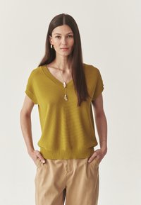 Pull en tricot jaune moutarde avec un col en V et des manches courtes, associé à un pantalon beige. Tissu texturé avec un ourlet côtelé.
