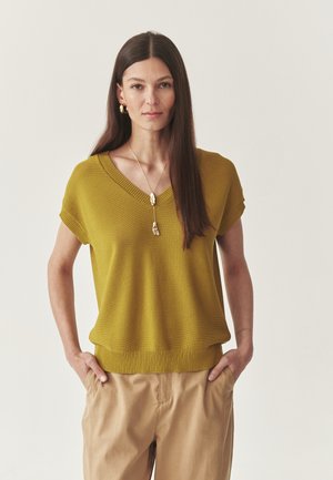 Senfgelber Strickpullover mit V-Ausschnitt und kurzen Ärmeln, kombiniert mit beigen Hosen. Strukturiertes Material mit geripptem Saum.