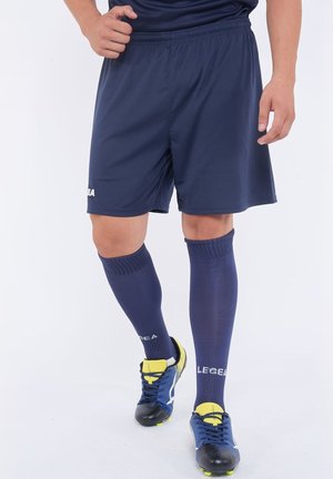 Personne portant un short de sport bleu marine, des chaussettes longues assorties avec un logo, et des crampons bleus et jaunes, debout sur un fond uni.