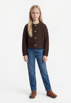 Noor tüdruk pikkade blondide juustega, kellel on pruun nööpidega kardigan, sinised teksad ja pruunid pahkluu saapad, seisab valgel taustal.