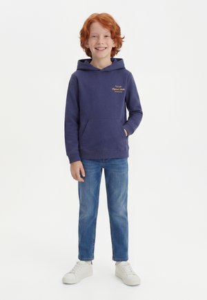 Garçon roux souriant, portant un sweat à capuche bleu marine, un jean bleu et des baskets blanches, debout avec une main dans la poche.