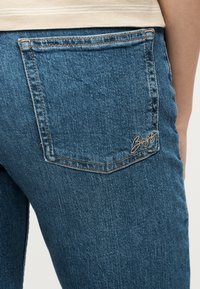 Κοντινό πλάνο πίσω τσέπης μπλε τζιν με ορατά ράμματα και κεντημένη λεπτομέρεια επωνυμίας στο δεξί ισχίο.