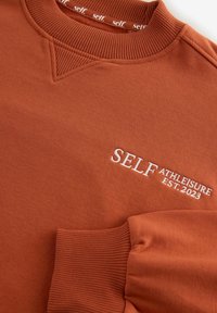 Ruskea huppari, jossa on ribbireunus kauluksessa ja hihoissa, etuosassa valkoiset brodeeratut tekstit "SELF ATHLEISURE EST. 2023".