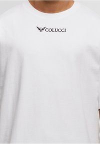 Tricou alb cu logo-ul Colucci de culoare neagră și un emblema stilizată cu aripi, centrată sub decolteu, pe partea superioară a toracelui unei persoane.
