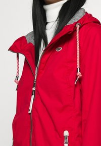 Červená nepromokavá bunda se šedou kapucí, zdobená stříbrnými zipy, pletenými stahovacími šňůrkami a malým logem na hrudi. Hladká textura.