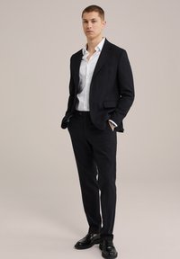 Traje negro con chaqueta ajustada y pantalones slim-fit, combinado con una camisa blanca ceñida y mocasines negros. Tejido suave, líneas limpias.