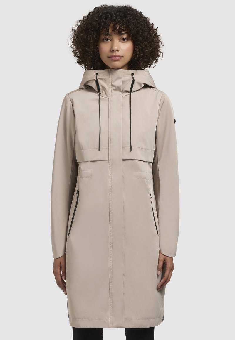 Beige vattentät kappa med huva, som har en dragkedjestängning, två framfickor, svarta detaljer och en medellång design. Slen struktur.