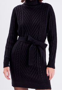 Robe en tricot noire avec col montant, texture côtelée, câbles verticaux et ceinture nouée à la taille. Longueur mi-cuisse avec manches longues.