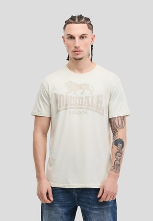 Homme aux cheveux tressés portant un t-shirt Lonsdale London beige clair et un jean bleu, montrant des tatouages sur son avant-bras gauche.