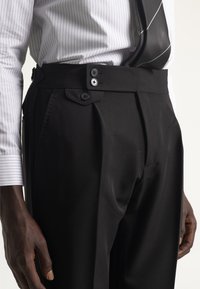 Hombre que lleva pantalones de tiro alto de cuadros negros con cintura de doble botón, combinados con una camisa blanca a rayas finas y una corbata de rayas negras.