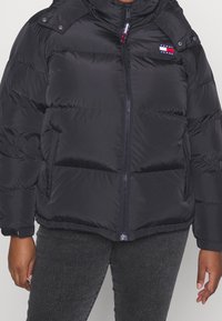 Giacca a vento nera con zip frontale, collo alto e dettagli con bottoni a pressione. Presenta un logo sul lato sinistro del petto e un tessuto testurizzato.