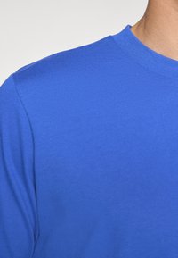 Tricou cu mânecă scurtă și guler rotund, realizat din bumbac albastru vibrant. Textură netedă, cu un design mulat, având bandă de gât și cusături pe umeri cu dungi.