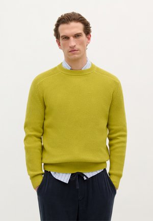 Geelgroene geribde trui met een ronde hals, lange mouwen en een gestructureerde stof. G gedragen over een blauw-wit gestreept overhemd met kraag.