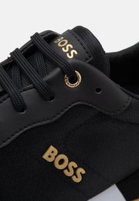 Svart sneaker med en texturerad tygovandel, släta läderdetaljer, guld-logotyp och runda snören. Har en kontrasterande vit sula.